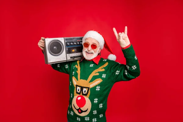 Photo of elderly funny santa man holding big vintage tape recorder chilling at party show horns wear trendy sun specs x-mas ugly ornament sweater cap isolated red background — Stock Photo, Image