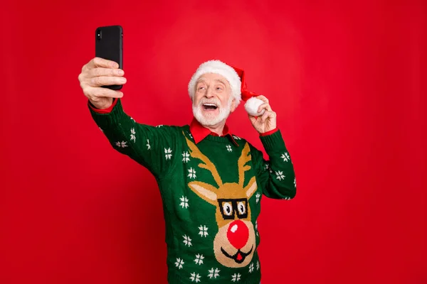 Bild av Santa man som håller telefon gör selfies för sociala nätverk x-mas blogg bära elegant x-mas ful prydnad tröja mössa isolerad röd bakgrund — Stockfoto