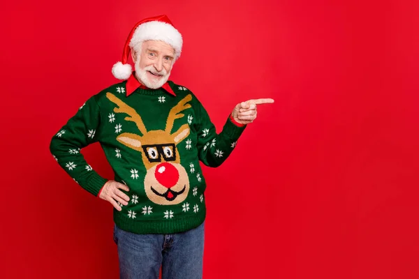 Photo of senior man indicating finger to empty space advising shocking low december prices wear x-mas ugly ornament sweater santa cap isolated red background — Stock Photo, Image