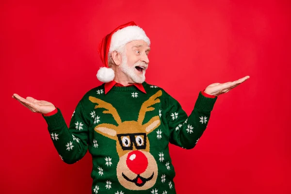 Photo of senior man holding sale products on open palms advising low holiday prices wear x-mas ugly ornament sweater and santa cap isolated red background — Stock Photo, Image