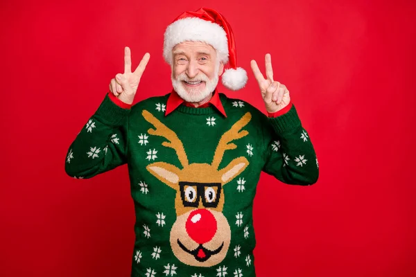 Portrait of positive cheerful old man in santa claus headwear have christmas x-mas party wear trendy reindeer decor pattern jumper make v-signs isolated over red color background — Stock Photo, Image