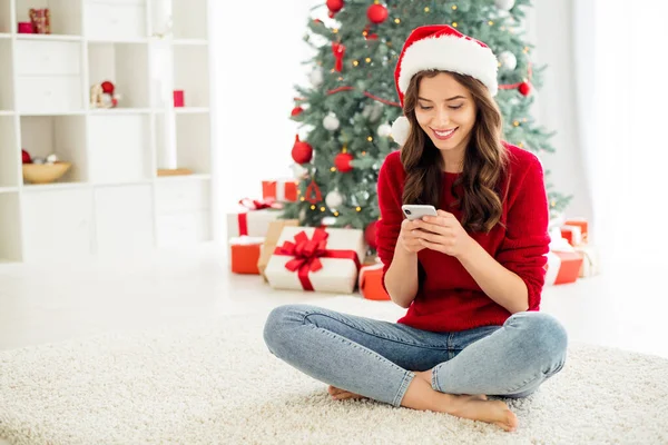 Çekici, çekici ve güzel bir kız arkadaşın kırmızı Noel Baba şapkası başlığı giyerken akıllı telefonuyla sohbet ettiği bir fotoğraf. — Stok fotoğraf