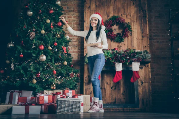 Foto de corpo inteiro de positivo alegre menina de cabelo marrom se preparar para x-mas tempo festa celebração decorar árvore de Natal com bolas brinquedos em casa com decoração do ano novo enfeites guirlandas dentro de casa — Fotografia de Stock