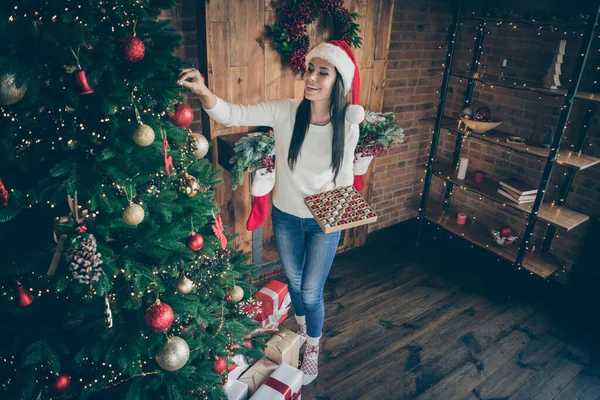원문 기사보기 산타클로스 모자를 쓴 긍정적 인 소녀의 높은 각도로 빛나는 소녀의 사진 이 크리스마스 트리에 새빨간 바우 블에 걸려 있다. — 스톡 사진