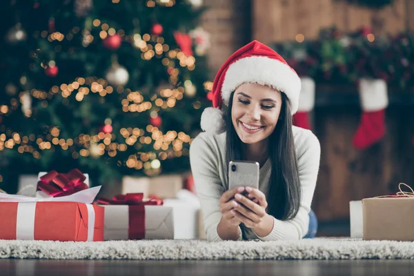 Close up photo of positive girl in Santa Claus hat lie on carpet use smartphone send christmas gratulacje szukaj rabaty w domu z x-mas komfort atmosfera w pomieszczeniach — Zdjęcie stockowe