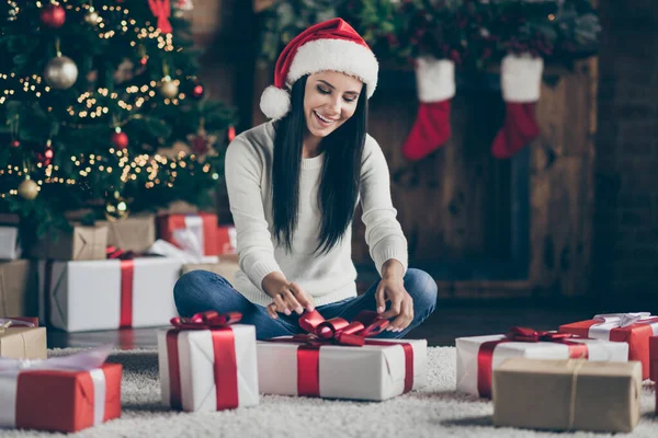 Πλήρες μέγεθος φωτογραφία του χαρούμενου θετική κορίτσι κάθονται πάτωμα στο σπίτι της με στολίδια newyear γιρλάντες προετοιμάσει το δώρο των ονείρων για τον άντρα της για τη νύχτα των Χριστουγέννων γιορτή κόμμα φορούν santa claus καπέλο σε εσωτερικούς χώρους — Φωτογραφία Αρχείου
