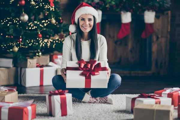 Full length photo of positive girl wear santa claus hat hold big giftbox get on Christmas night enjoy newyear tradition sit on floor carpet in house with celebratic x-mas illumination indoor — Φωτογραφία Αρχείου