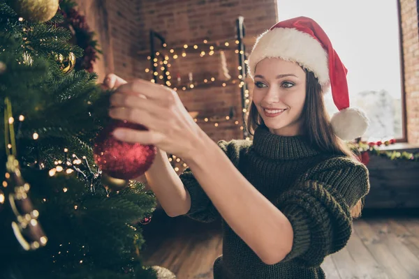 Närbild foto av positiv glad flicka i Santa claus mössa stickad tröja njuta av jul firande hänga röd stor grannlåt på vintergröna träd i huset med nyår dekoration inomhus — Stockfoto