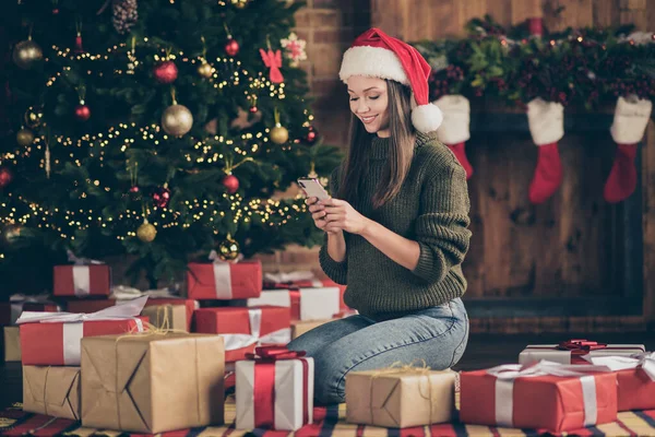 Volledige foto van positief meisje in de kerstman hoed gebruik maken van mobiele telefoon te lezen gefeliciteerd zoeken traditie x-mas kortingen zitten gezellige vloer geruite deken surround giftbox in huis binnen — Stockfoto