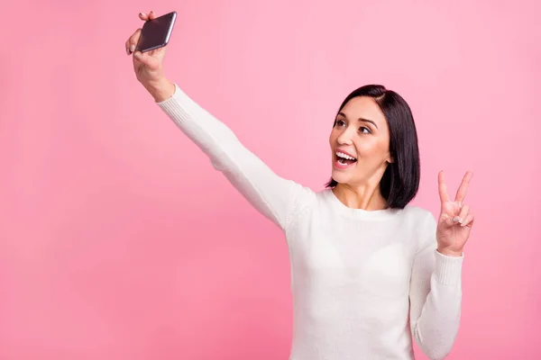 Фото удивительной леди, держащей телефон в руках, делая селфи, показывающие V-знак символ носить белый пуловер изолированный розовый яркий цвет фона — стоковое фото