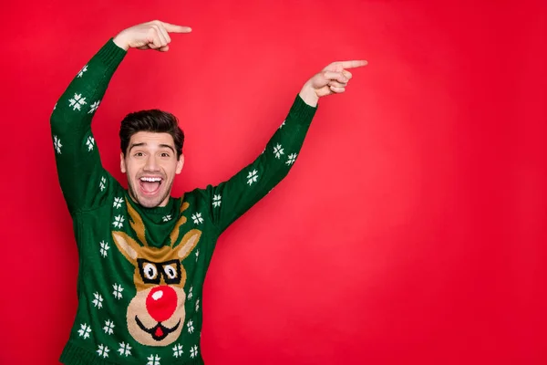 Foto von emotionalen gutaussehenden Kerl zeigt Finger auf Weihnachtsverkauf Banner tragen trendige gestrickte grüne Pullover mit Hirsch isoliert roten Farbhintergrund — Stockfoto