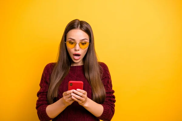 Портрет впечатленной девушки в очках, читающей информацию со своего гаджета, одетой в бордовый пуловер, изолированный на жёлтом фоне — стоковое фото