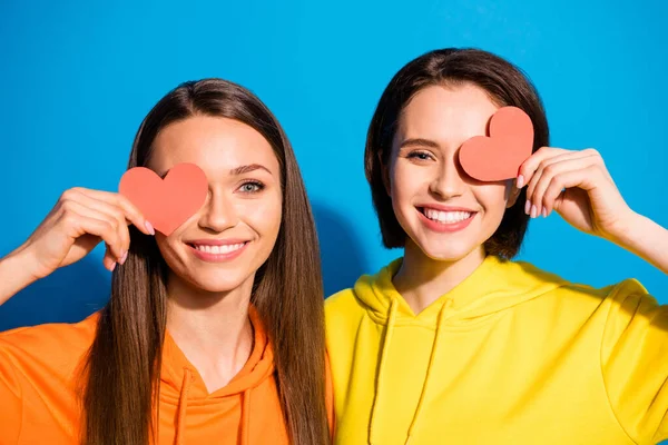 Foto de dos bonitas dos damas cogidas de la mano pequeños corazones de papel rojo que ocultan los ojos usan sudaderas con capucha amarillas anaranjadas aisladas color azul fondo — Foto de Stock