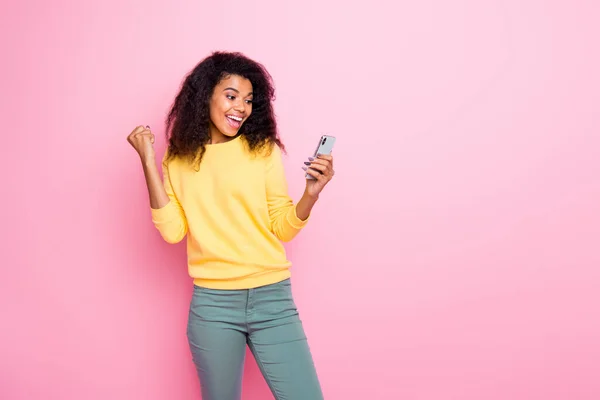 Retrato de encantado funky loca africana chica blogger utilizar su teléfono celular recibir notificación comentarios comentarios levantar puños gritar sí usar pantalones suéter amarillo aislado sobre pastel color fondo — Foto de Stock