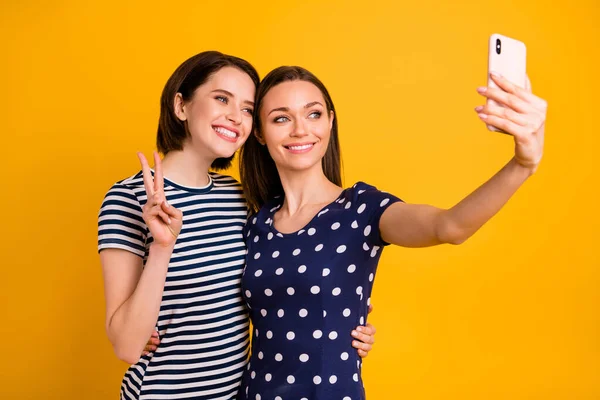 Photo of two amazing ladies holding telephone making selfies showing v-sign symbols wear summer casual trendy t-shirts isolated yellow background — Stock Photo, Image