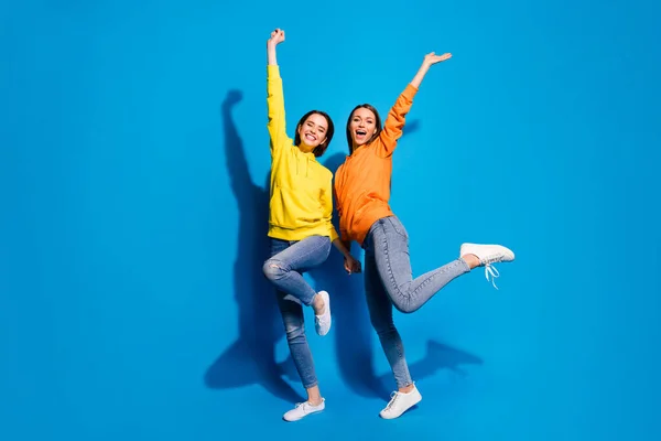 Полнометражное фото двух милых дам, поднимающих руки, путешествующих за границу, в повседневных ярких толстовках и джинсах с голубым цветом фона. — стоковое фото