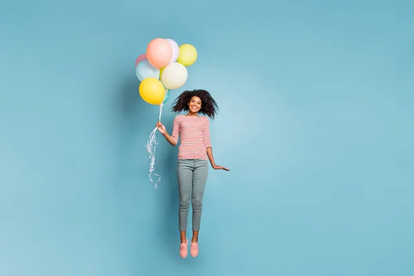 Zdjęcie kręcone faliste wesoły modny ładna dziewczyna nosi spodnie pasiasty t-shirt lecący w górę trzymając balony izolowane nad niebieskim pastelowym tle kolor — Zdjęcie stockowe