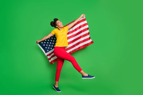 全长照片，迷人的深色皮肤女士庆祝独立美国日，游行沼泽地上挂着国旗，身穿休闲黄T恤红色裤子，衬托着孤立的绿色背景 — 图库照片