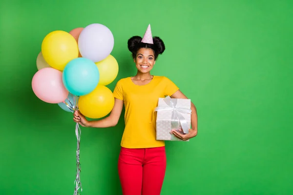 Foto di bella signora pelle scura in possesso di molti palloncini d'aria e grande giftbox è venuto alla festa di compleanno indossare casual giallo t-shirt pantaloni rossi tappo di carta isolato sfondo verde — Foto Stock