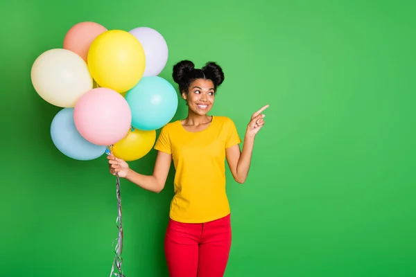 Photo of pretty dark skin lady holding many air balloons directing finger empty space on sale prices wear casual yellow t-shirt red pants isolated green background
