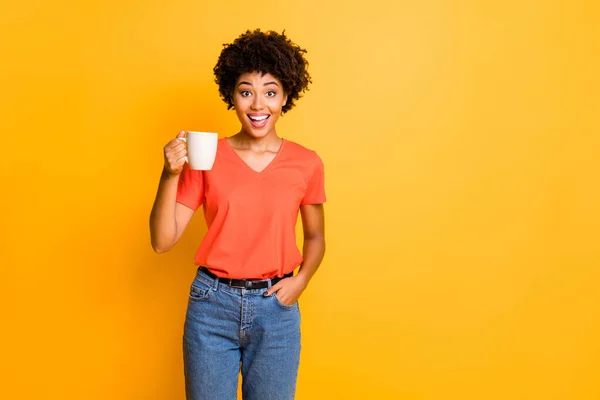 Sarı renkli arka planda izole edilmiş çay bardağı tutan turuncu t-shirt kot pantolon giyen sevimli, neşeli, çekici kız arkadaşın fotoğrafı. — Stok fotoğraf