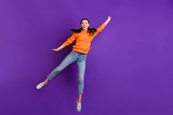 Teljes méretű fotó a pozitív vidám tini lány hold hand jump fogás neki repülő napernyő a szeles időjárás viselni narancssárga sportos ruha farmer fehér cipő elszigetelt felett lila színű háttér — Stock Fotó