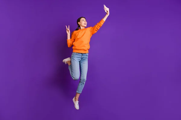 Volledige foto van positieve vrolijke jeugd tiener meisje met paardenstaarten hebben reis maken selfie maken v-tekens dragen oranje sportieve dragen jeans sneakers geïsoleerde violet paarse kleur achtergrond — Stockfoto