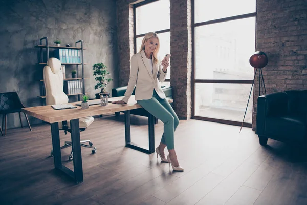 Full size foto di fiduciosa focalizzata leader di mezza età donna d'affari sedersi sul tavolo di legno in ufficio utilizzare la sua chat smartphone con i suoi clienti partner di workstation indossare tacchi alti — Foto Stock