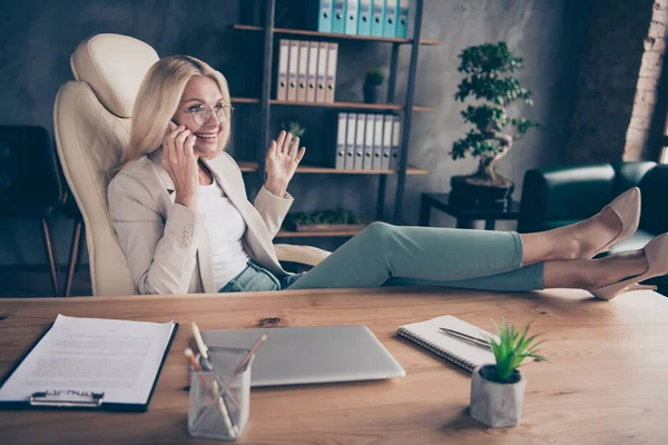 Foto de empreendedora positiva alegre tendo terminado de trabalhar em seu projeto e dando ao luxo de falar por telefone com as pernas na mesa — Fotografia de Stock