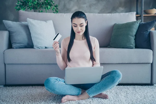Foto de una señora adicta sosteniendo un cuaderno en las rodillas que va a pagar en línea con la ayuda de la tarjeta de crédito sentado piso en la alfombra cómoda usando jersey y jeans en el apartamento — Foto de Stock