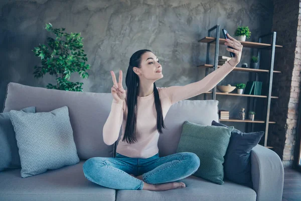 Photo de jolie dame avec téléphone dans les mains faisant selfies montrant le symbole de v-signe assis confortablement sur le canapé portant pull et jeans homey dans l'appartement — Photo