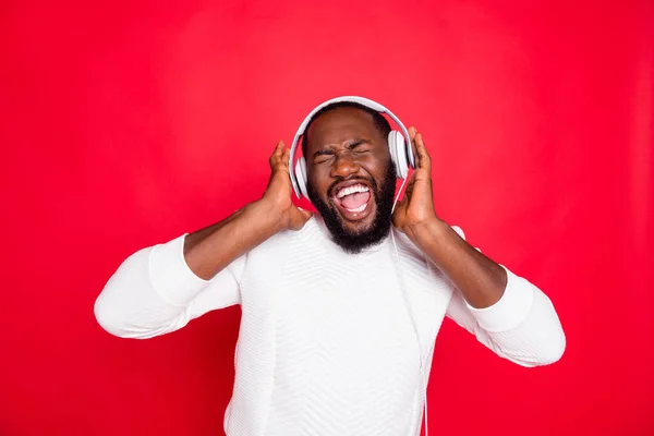 Foto de engraçado homem de pele escura ouvindo playlist favorito em earflaps loucos cantando legal faixa desgaste branco malha suéter isolado fundo vermelho — Fotografia de Stock