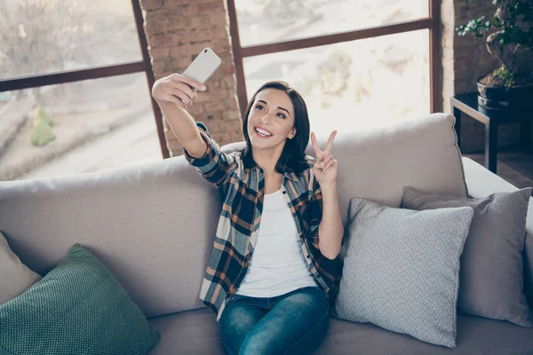 Foto de la encantadora dama sosteniendo el teléfono haciendo selfies para el blog mostrando el símbolo del signo v sentado cómodo sofá con camisa a cuadros casual y apartamento de vaqueros en el interior — Foto de Stock