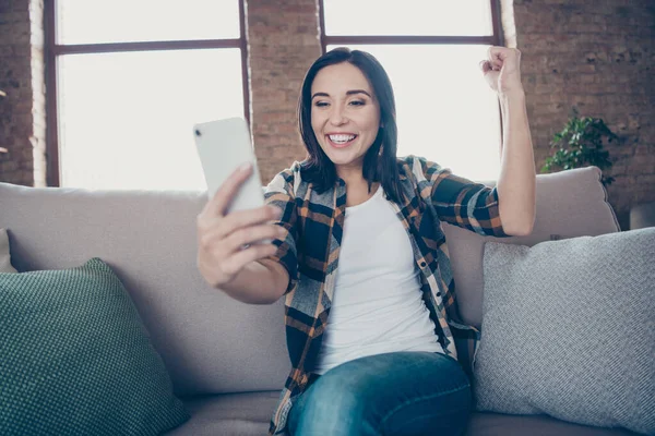 Фото красавицы, держащей телефон, читающей лучшие новости о стартап-проекте, поднимающей кулак, сидящий на уютном диване в повседневной одежде — стоковое фото