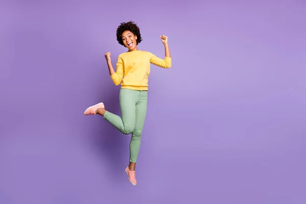 Volledige lengte foto van opgewonden afro-Amerikaanse meisje horen nieuws geluk herfst overwinning onder de indruk gil wow sprong raise vuisten dragen geel pullover groene broek geïsoleerde violet kleur achtergrond — Stockfoto