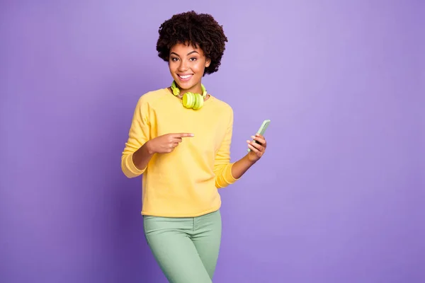 Foto de la onda de moda elegante alegre alegre alegre linda novia dulce con pantalones verdes pantalones suéter amarillo sonriendo toothily apuntando a su teléfono con auriculares aislados sobre puple — Foto de Stock