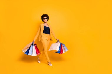 Full body photo of positive cheerful afro american girl on travel trip go shopping found sales discounts hold bags wear vogue style outfit sunglass pants high-heels isolated yellow color background clipart