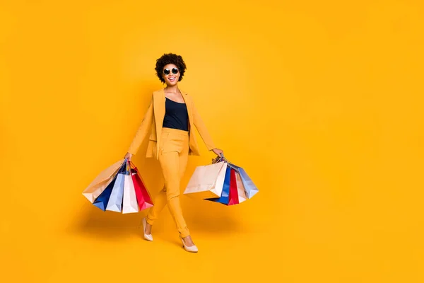 Teljes test fotó a pozitív vidám afro amerikai lány az utazás menni vásárlás talált értékesítési kedvezmények tartsa táskák viselet divatos stílus ruha napszemüveg nadrág magassarkú elszigetelt sárga háttér — Stock Fotó