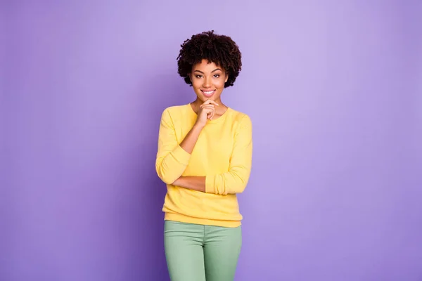 Foto de la alegre chica linda desconcertada con ropa amarilla posando frente a la cámara tocando su barbilla sonriendo con un rayo de dientes aislado sobre el fondo de color pastel púrpura — Foto de Stock