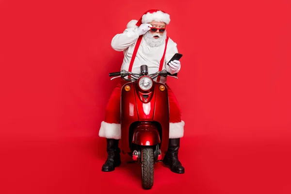 Portret ładne atrakcyjne brodaty szokujące śmieszne funky Santa noszenie czapki kapelusz siedzi na motocyklu za pomocą wiadomości SMS czytania komórek izolowane na jasny żywy połysk żywy czerwony kolor tła — Zdjęcie stockowe
