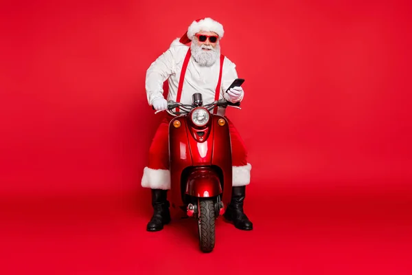 Portrait of nice attractive bearded cheerful cheery Santa wearing cap hat sitting on motor bike using cell taking orders isolated over bright vivid shine vibrant red color background — Stock Photo, Image