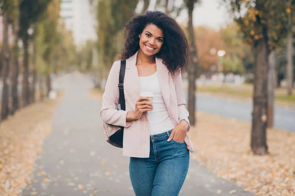 Foto de encantadora pele escura encaracolado senhora caminhando parque depois de palestras da faculdade beber takeaway quente café segurar mochila quente queda temporada desgaste casaco jeans ao ar livre — Fotografia de Stock