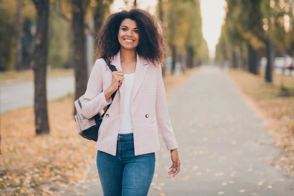 Photo of amazing pretty dark skin lady toothy smiling spędza wolny czas spacerując park po wykładach uniwersyteckich na zewnątrz gospodarstwa plecak jesień sezon noszenia dżinsy kurtka — Zdjęcie stockowe