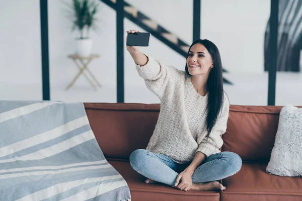 Portrait de fille gaie positive tenir téléphone portable prendre selfie photo assis sur divan dans la maison appartement moderne sentir contenu porter pull tricoté jeans en denim — Photo