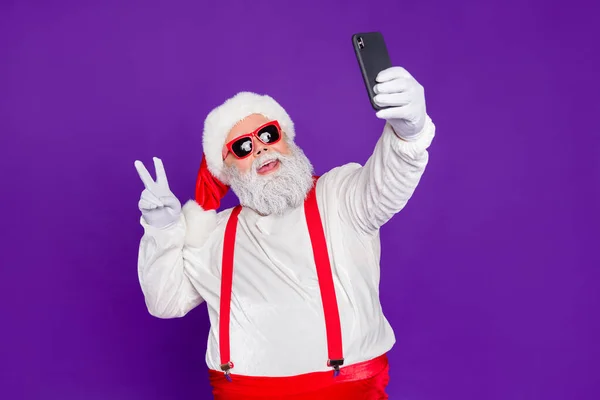 Portrait of his he nice funny cheerful cheery bearded Santa taking making selfie showing v-sign travel trip blogging isolated over bright vivid shine vibrant violet lilac background — Stockfoto