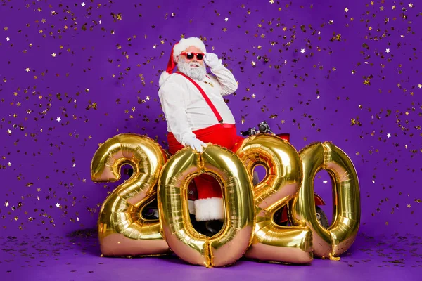 Хо-хо-хо. Фотографія повного тіла Санти чоловік сидить на мотоциклі повітряних кульок нового року 2020 року, говорячи, що привітання носять сонячні окуляри костюм ізольований фіолетовий фон — стокове фото
