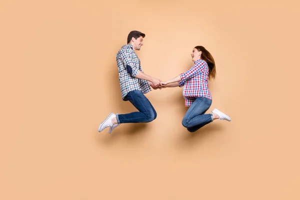 Full längd profil foto av två personer galen kvinna kille hoppar högt hålla händerna glada lekfull humör bära casual pläd jeans kläder isolerad beige färg bakgrund — Stockfoto