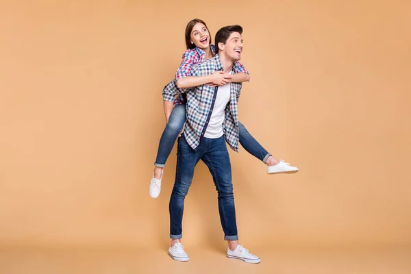 Tamanho do corpo de comprimento total virou foto de alegre positivo bonito casal branco bonito vestindo jeans ganga com ela tendo montado suas costas e ele carregando-a isolada sobre fundo cor pastel bege — Fotografia de Stock