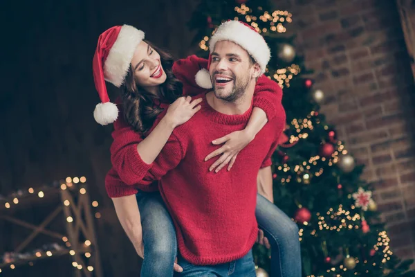 Foto de pareja funky pasando la víspera de x-mas en la sala de luces de guirnalda decorada llevando a cuestas celebración humor desgaste pullovers rojos y sombreros de santa en el interior —  Fotos de Stock