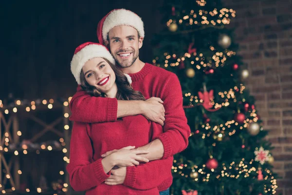 Zdjęcie uroczej pary spędzającej Wigilię Bożego Narodzenia w urządzonym pokoju z lampkami do pończoch stojący świnka w pobliżu x-mas drzewa w pomieszczeniach nosić czerwone pulowery i czapki Mikołaja — Zdjęcie stockowe